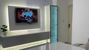 Uma sala de estar moderna com televisão de tela plana na parede, uma mesa de centro branca, um sofá, um aparador de vidro e uma porta.