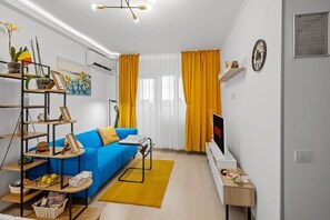 Ett vardagsrum med en blå soffa, en tv, en gul matta och en hylla för växter.