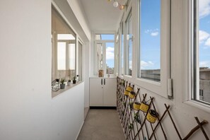 Ein schmaler Korridor mit weißen Wänden, einem gefliesten Boden und großen Fenstern an einer Seite.