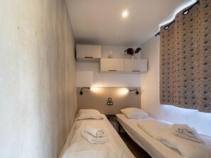 A hotel room with two single beds, a small bedside table, a wall-mounted shelf with a vase, and a patterned curtain. - Nice Family Caravan in the Countryside (Kaprijke)