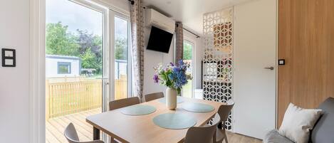 A modern room with a wooden dining table, chairs, a sofa, a TV, and a view of a wooden deck outside.