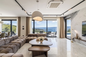 A modern living room with a sectional sofa, a round coffee table, and a large window offering a view of the outdoors. - Villa M&D, Luxury Sea View Villa with Pool & Sauna (Dugi Rat)
