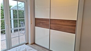 A room with a sliding door leading to an outdoor area, a bed with a white duvet, and a wardrobe with a wooden accent panel. - Deichhuus in Nessmersiel (Dornum)