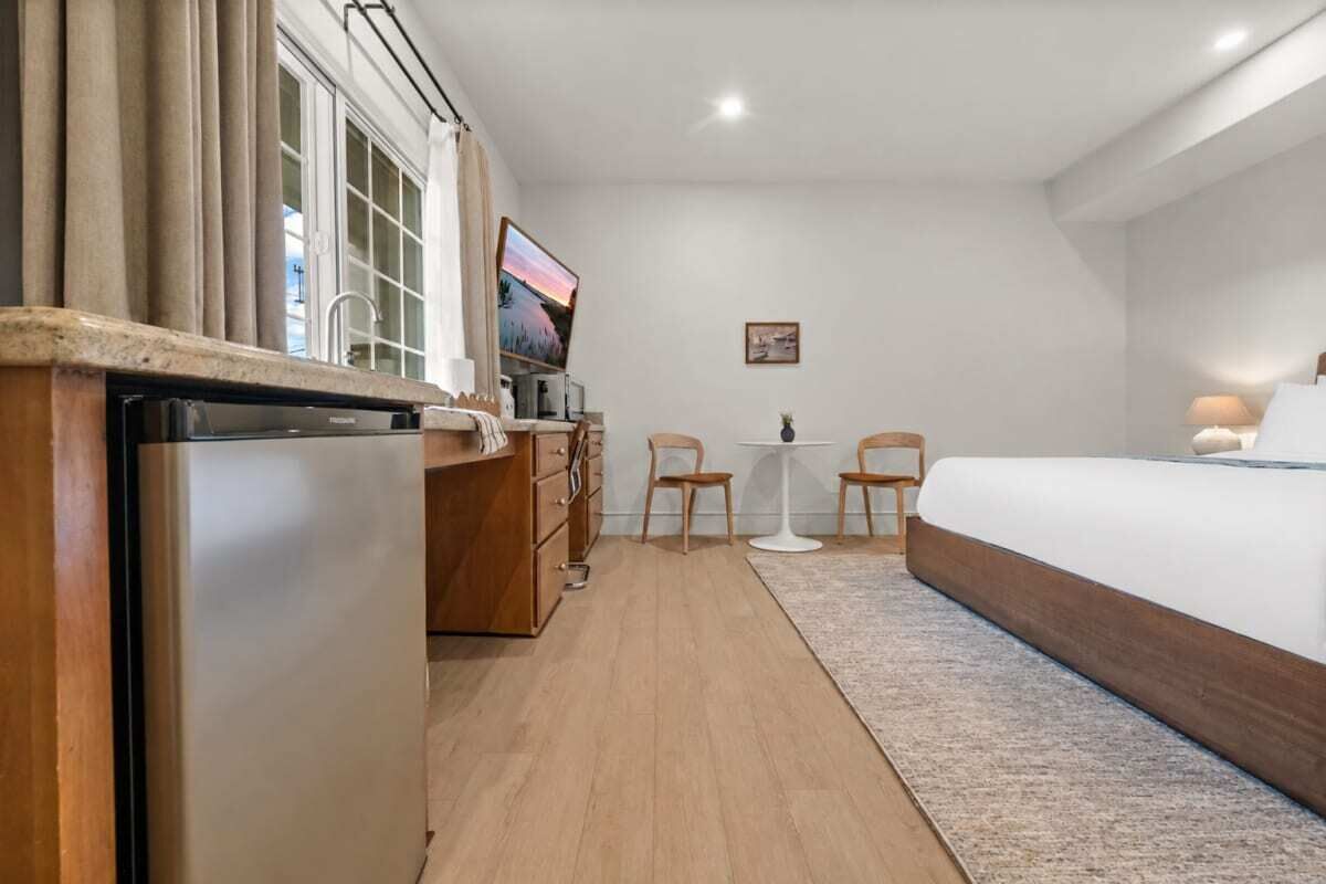 A hotel room with a bed, a small table, two chairs, a refrigerator, a TV, and a window with curtains.