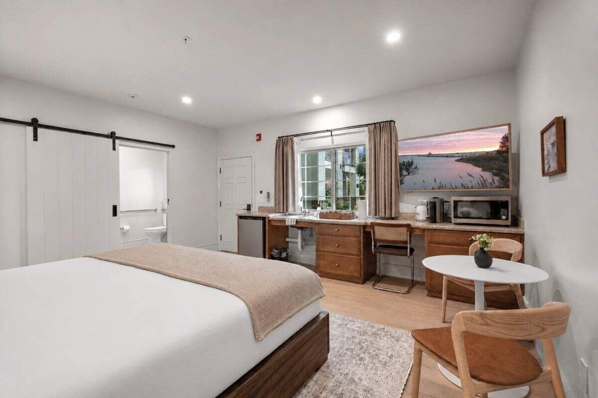 Een slaapkamer met een bed, een bureau, een stoel, een klein tafeltje, een raam met gordijnen, een schilderij en een kitchenette.