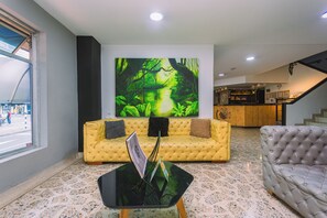 Un vestíbulo moderno con un sofá amarillo, un sofá gris, una mesa de centro de vidrio y un cuadro grande con temática de bosque en la pared.