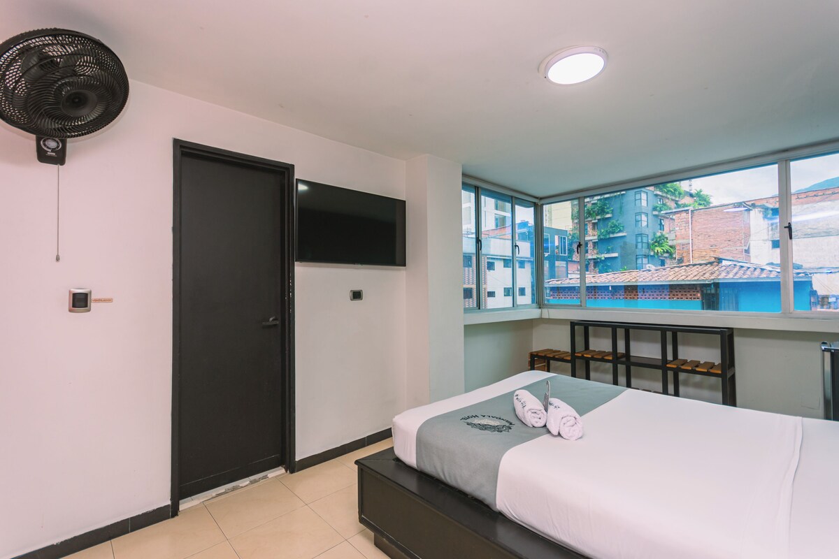 Un dormitorio con cama, televisor montado en la pared, ventana con vista a edificios y ventilador de techo.