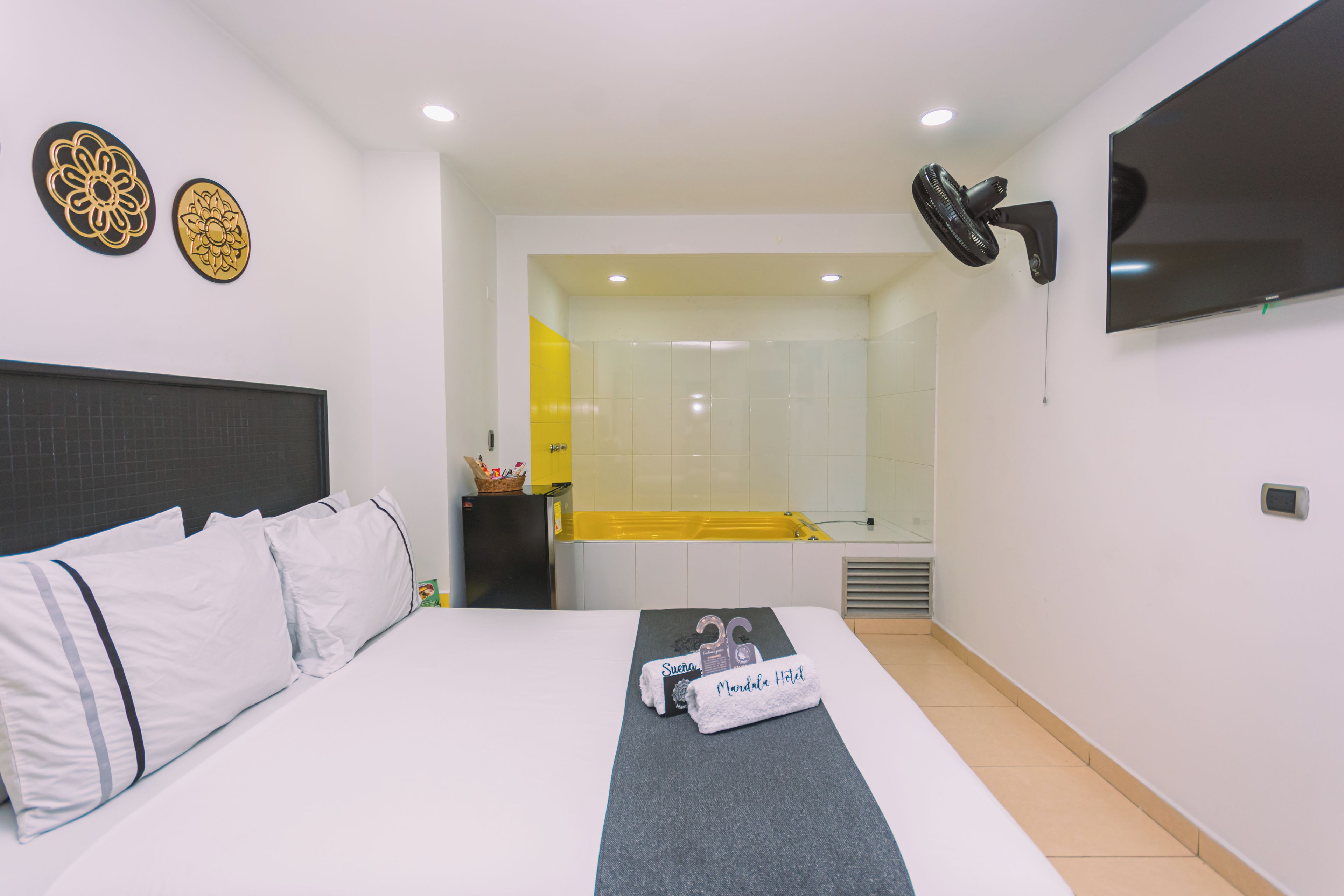 Habitación de hotel con cama, televisión, ventilador y baño visible a través de una puerta abierta.