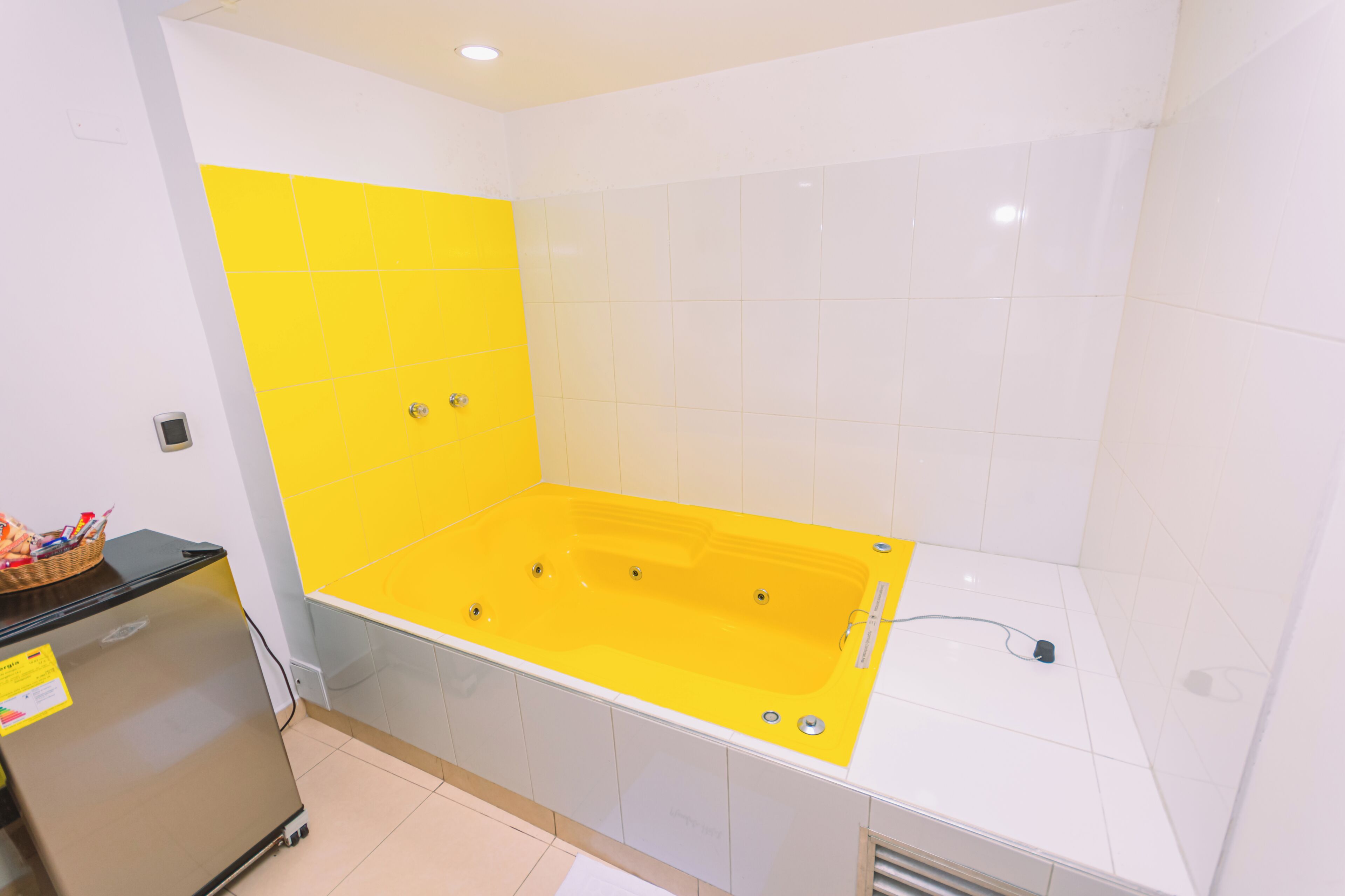 A bathroom with a yellow bathtub, white tiles, and a small refrigerator.