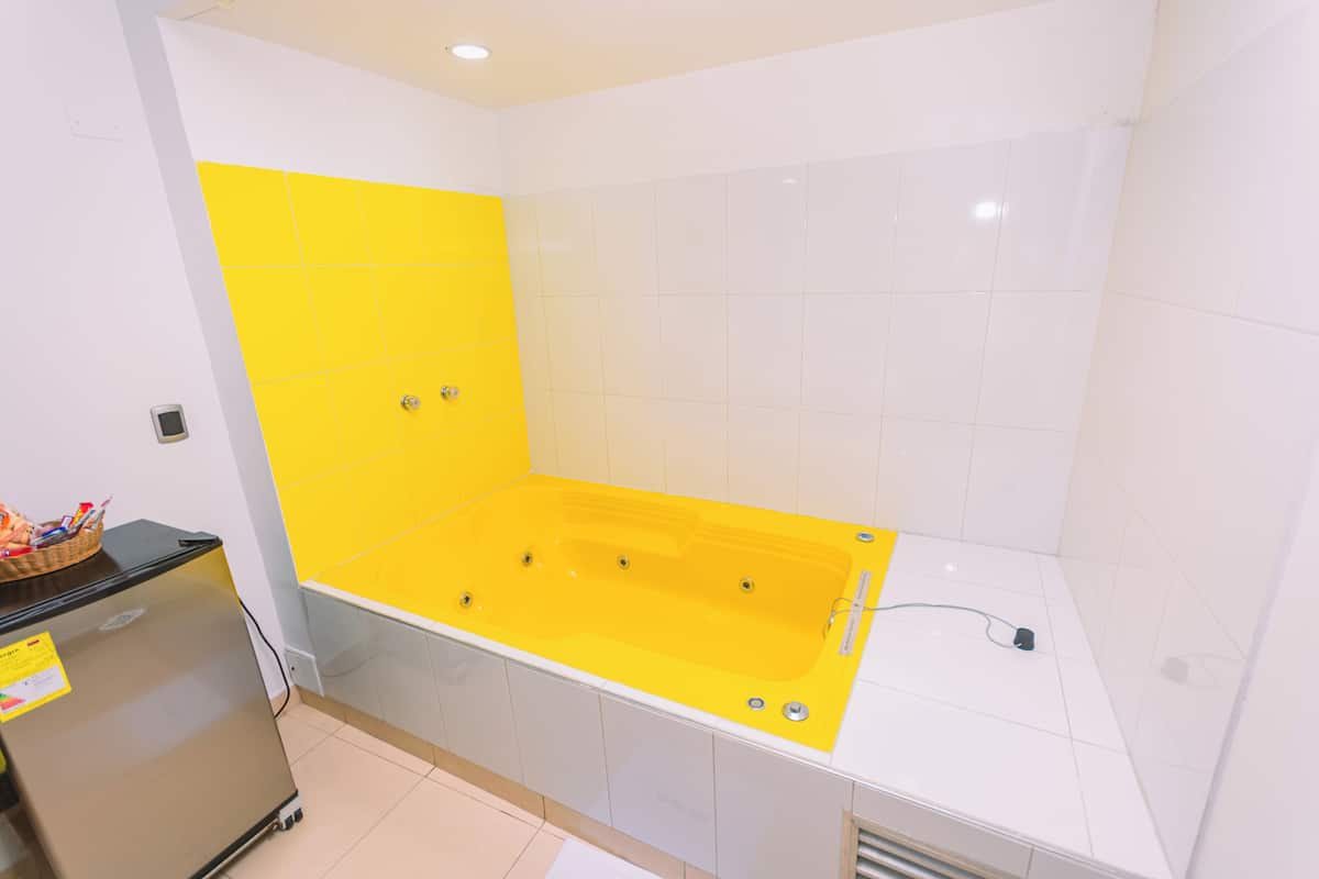 Un baño con una bañera amarilla, azulejos blancos y un refrigerador pequeño.