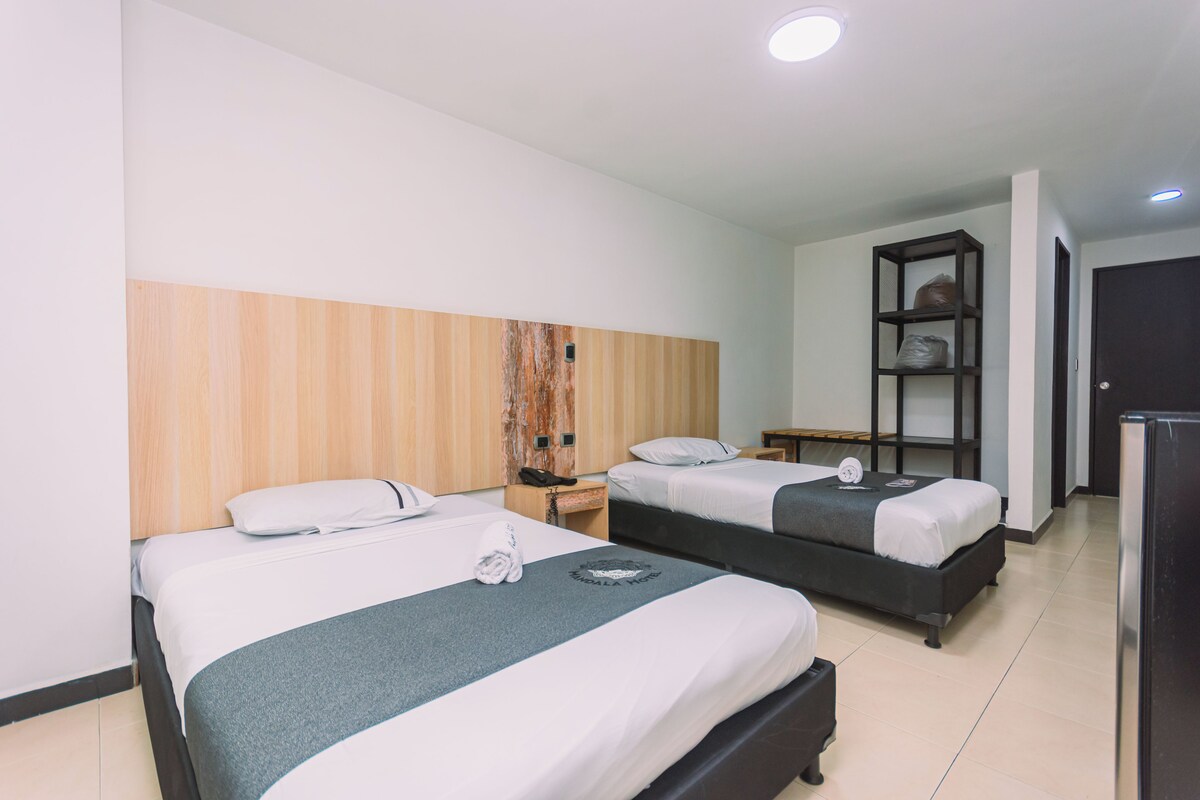 A hotel room with two beds, a wooden headboard, a small desk with a phone, and a shelf with items.