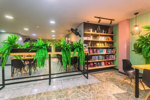 A cafe with a counter, chairs, a bookshelf with books and magazines, and potted plants. - Mandala Medellín Centro By Gyc Hotels (Medellín)