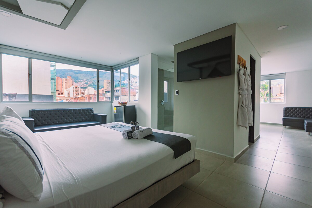 Una habitación de hotel moderna con una cama grande, zona de estar, televisor y vistas a la ciudad.
