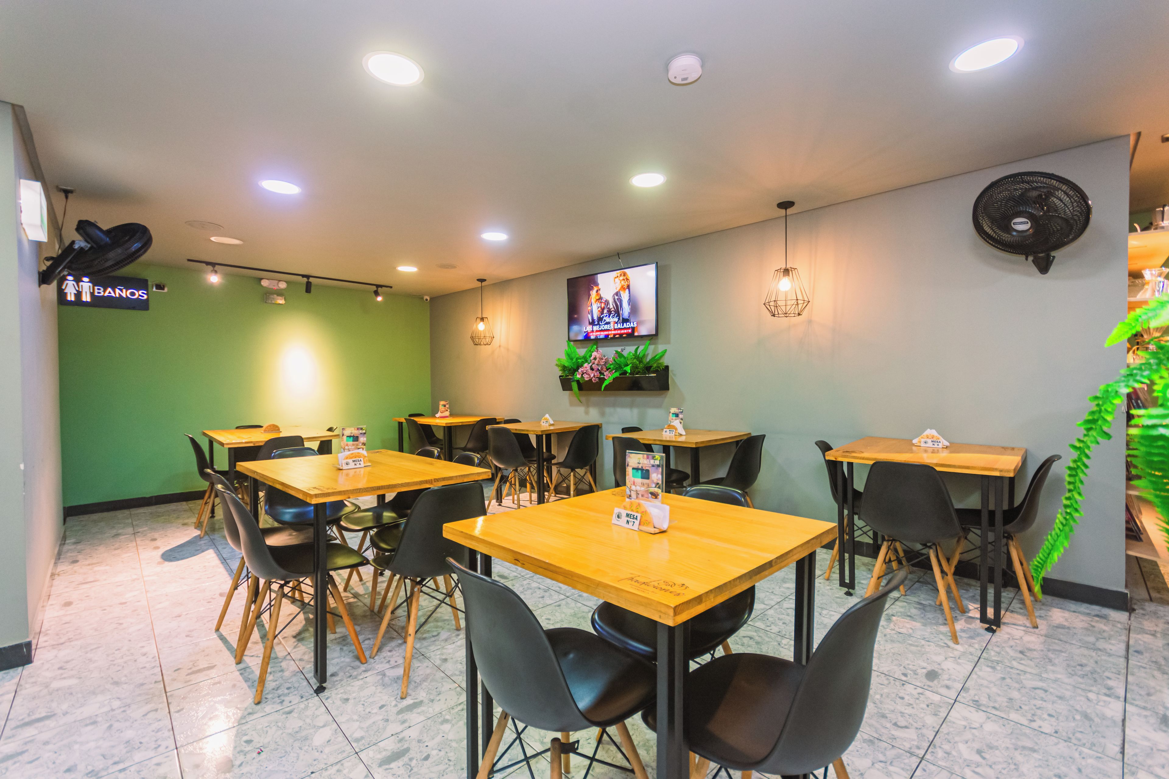 Interior de un restaurante con varias mesas y sillas, un televisor montado en la pared y un letrero que indica la ubicación de los baños.