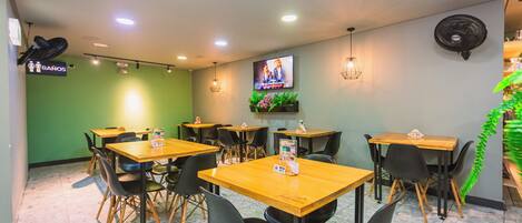 Interior de un restaurante con varias mesas y sillas, un televisor montado en la pared y un letrero que indica la ubicación de los baños.