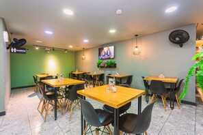 Interior de un restaurante con varias mesas y sillas, un televisor montado en la pared y un letrero que indica la ubicación de los baños.