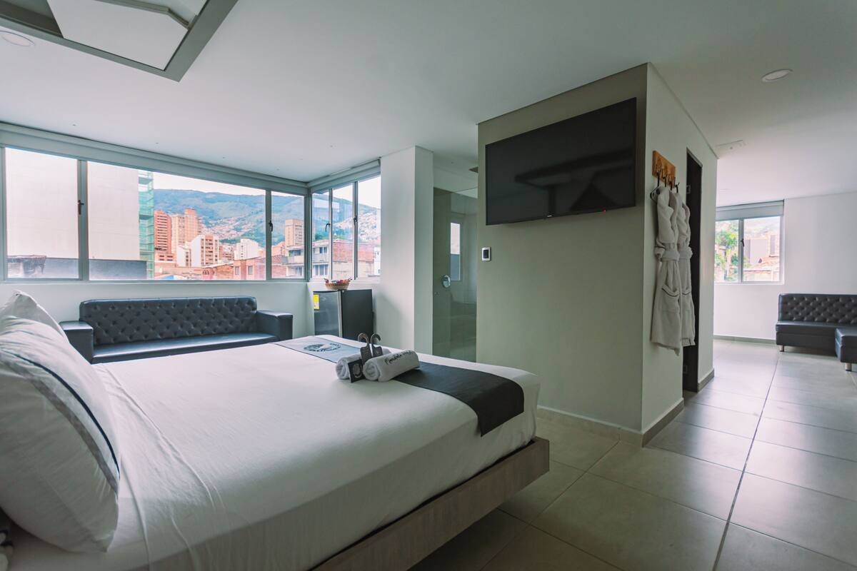 Una habitación de hotel moderna con una cama grande, zona de estar, televisor y vistas a la ciudad.