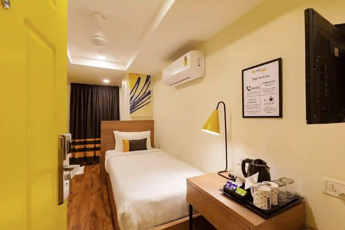 A hotel room with a bed, bedside table with a coffee maker, a lamp, an air conditioning unit, and a framed sign on the wall.