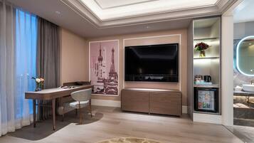 A modern hotel room with a flat-screen TV, a desk with a chair, a minibar, and a large window with curtains.