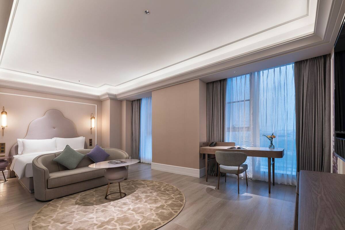 Una habitación de hotel moderna con cama, sofá, escritorio y un amplio ventanal con cortinas.