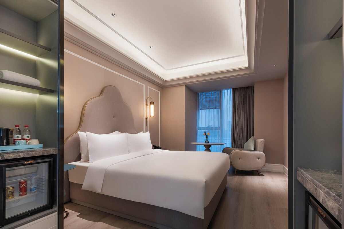 A modern hotel room with a large bed, a minibar, a small table with a vase, and a view of the city through a window.