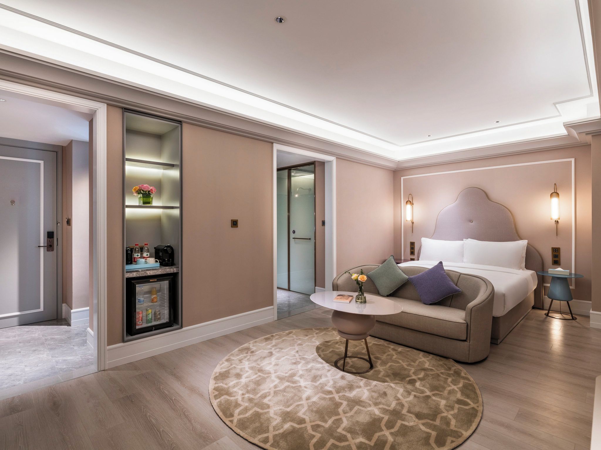 A modern hotel room with a bed, a small round table, a sofa, a coffee machine, and a minibar.