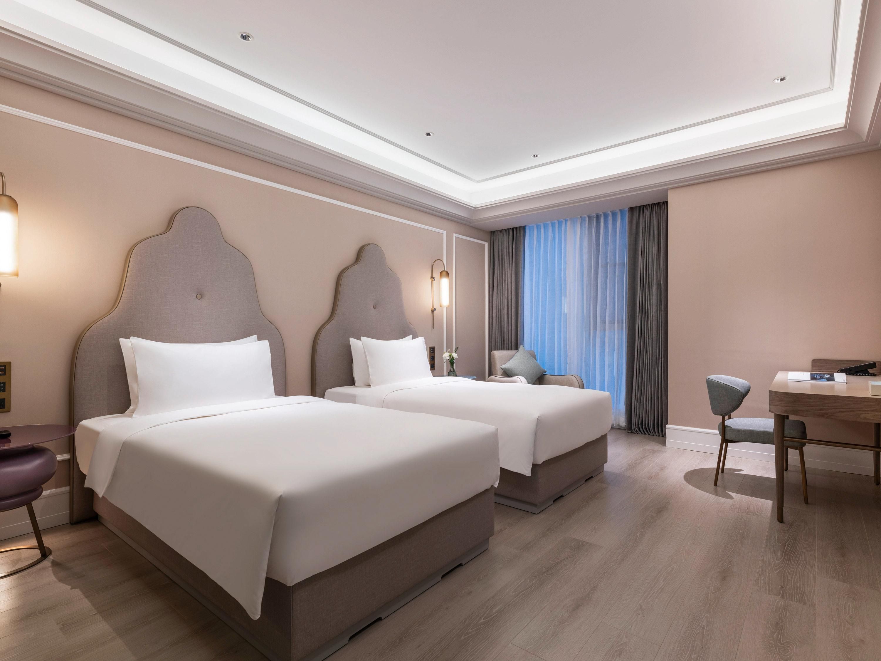A hotel room with two beds, a desk, a chair, and a large window with curtains.