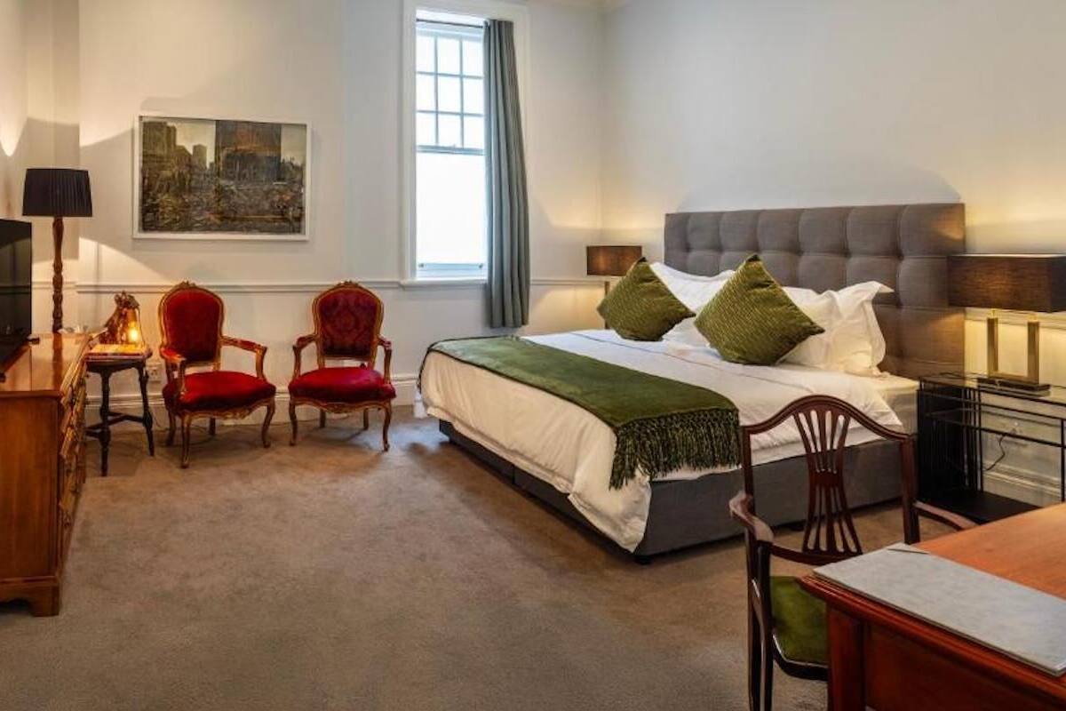 A bedroom with a bed, two chairs, a desk, a lamp, and a painting on the wall.