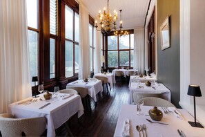 Un restaurant amb diverses taules, llums de cristall i grans finestres.