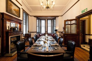 Un comedor formal con una larga mesa dispuesta para una comida, flanqueada por sillas de cuero, una chimenea y amplias ventanas.