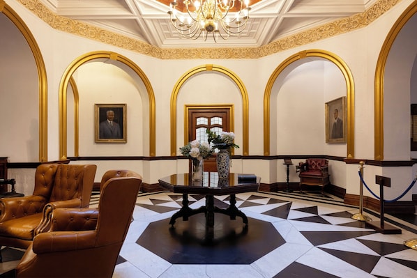Un vestíbulo con piso a cuadros, sillones de cuero, una mesa central con flores y retratos enmarcados en las paredes.