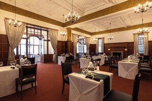 Comedor formal con varias mesas dispuestas para el servicio, ventanas amplias con cortinas y una lámpara clásica.