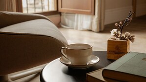 Ein gemütliches Zimmer mit einer Couch, einem runden Tisch mit Tasse und Büchern sowie einem Fenster mit Vorhängen.