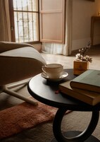 Una habitación acogedora con sofá, una mesa redonda con una taza y libros, y una ventana con cortinas.