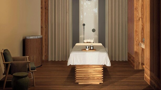 A room with a wooden floor, a bed with a white bedspread, a bedside table with a lamp, a chair, and a curtain.