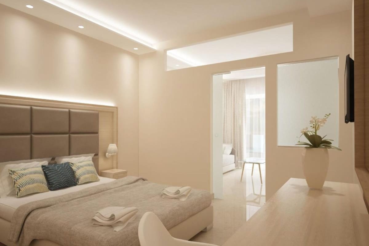 A modern hotel room with a bed, bedside table, lamp, and a view of the adjacent living area.