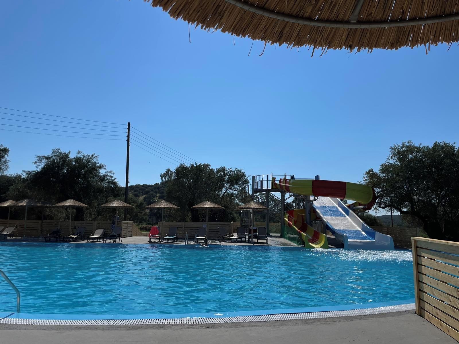 Una piscina con tobogán de agua, sillas de descanso y sombrillas de paja.