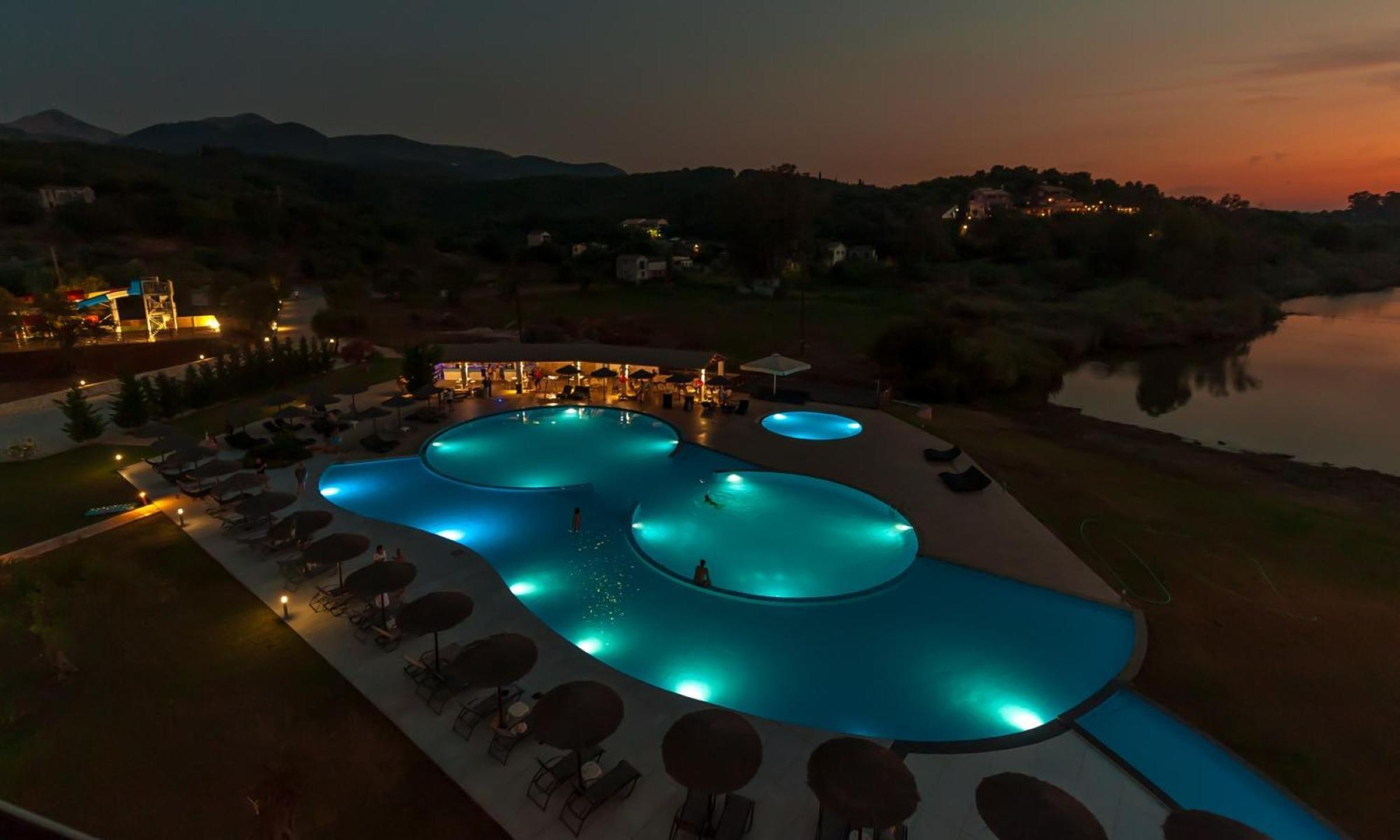 Una amplia piscina exterior con varias secciones, rodeada de sillas de descanso y sombrillas, con colinas y una masa de agua al fondo.