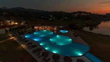 Una amplia piscina exterior con varias secciones, rodeada de sillas de descanso y sombrillas, con colinas y una masa de agua al fondo.