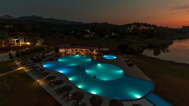 A large outdoor swimming pool with multiple sections, surrounded by lounge chairs and umbrellas, set against a backdrop of hills and a body of water. - Laguna Holiday Resort Corfu (Corfu)