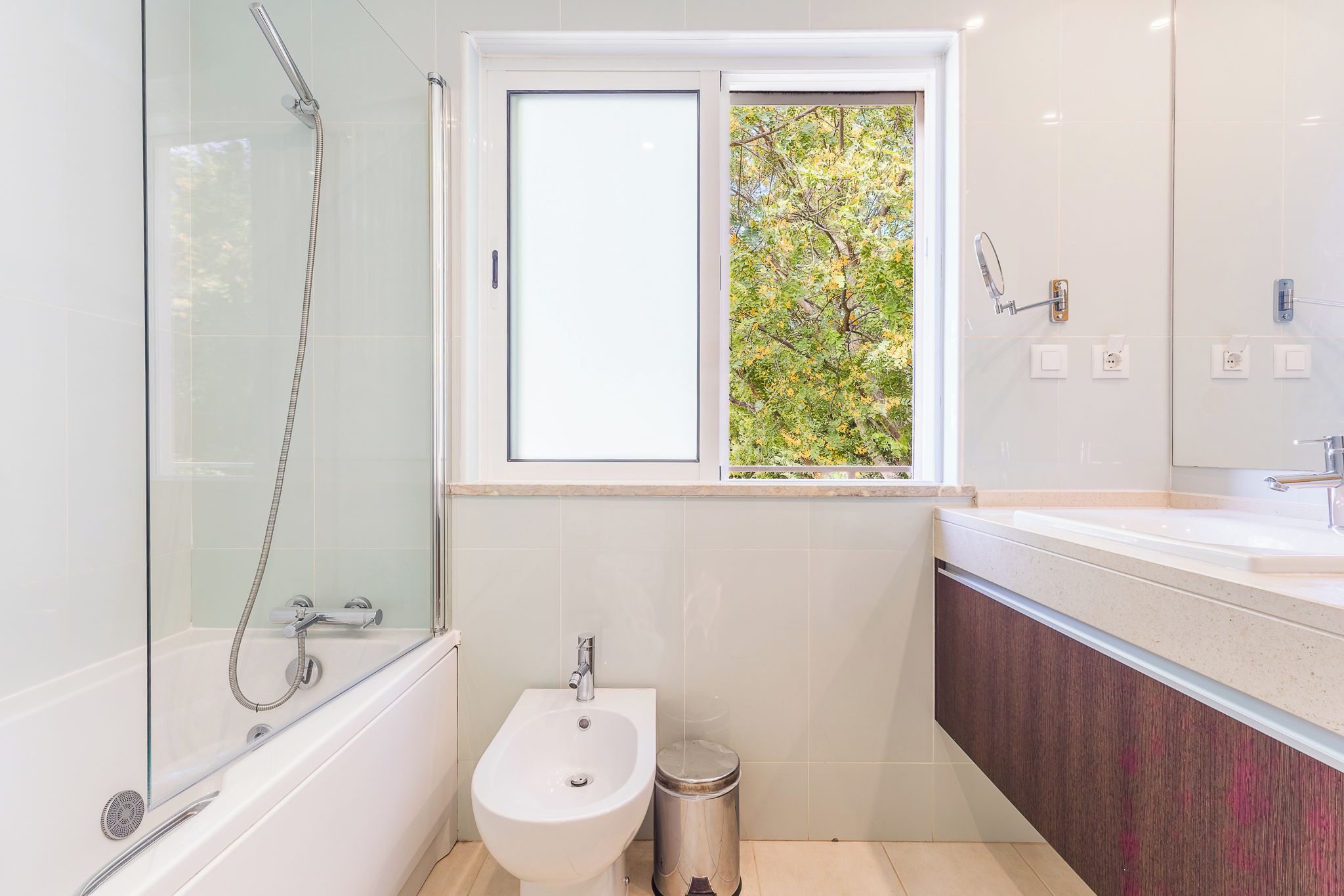 A modern bathroom with a glass-enclosed shower, a bidet, a sink, and a window with a view of greenery.