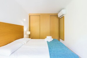 A hotel room with a wooden headboard, a bed with white linens, a blue throw blanket, a wall-mounted air conditioner, and a wooden wardrobe. - Lumina Villas (Lagoa)
