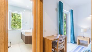 A modern room with a wooden desk, a chair, a small bedside table with a lamp, and a view of greenery through a window.