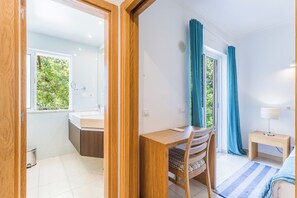 A modern room with a wooden desk, a chair, a small bedside table with a lamp, and a view of greenery through a window. - Lumina Villas (Lagoa)