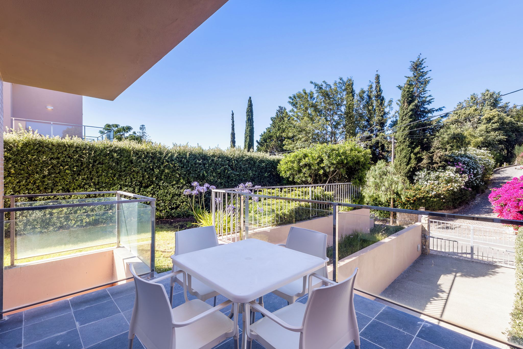 A balcony with a table and chairs, a glass railing, and a view of a garden with trees and flowers.