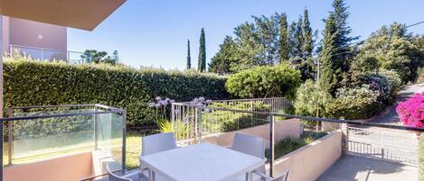 A balcony with a table and chairs, a glass railing, and a view of a garden with trees and flowers.