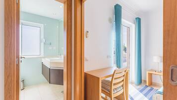 Una habitación con escritorio de madera, una silla, una cama, ventana con cortinas y baño con ducha y lavamanos.