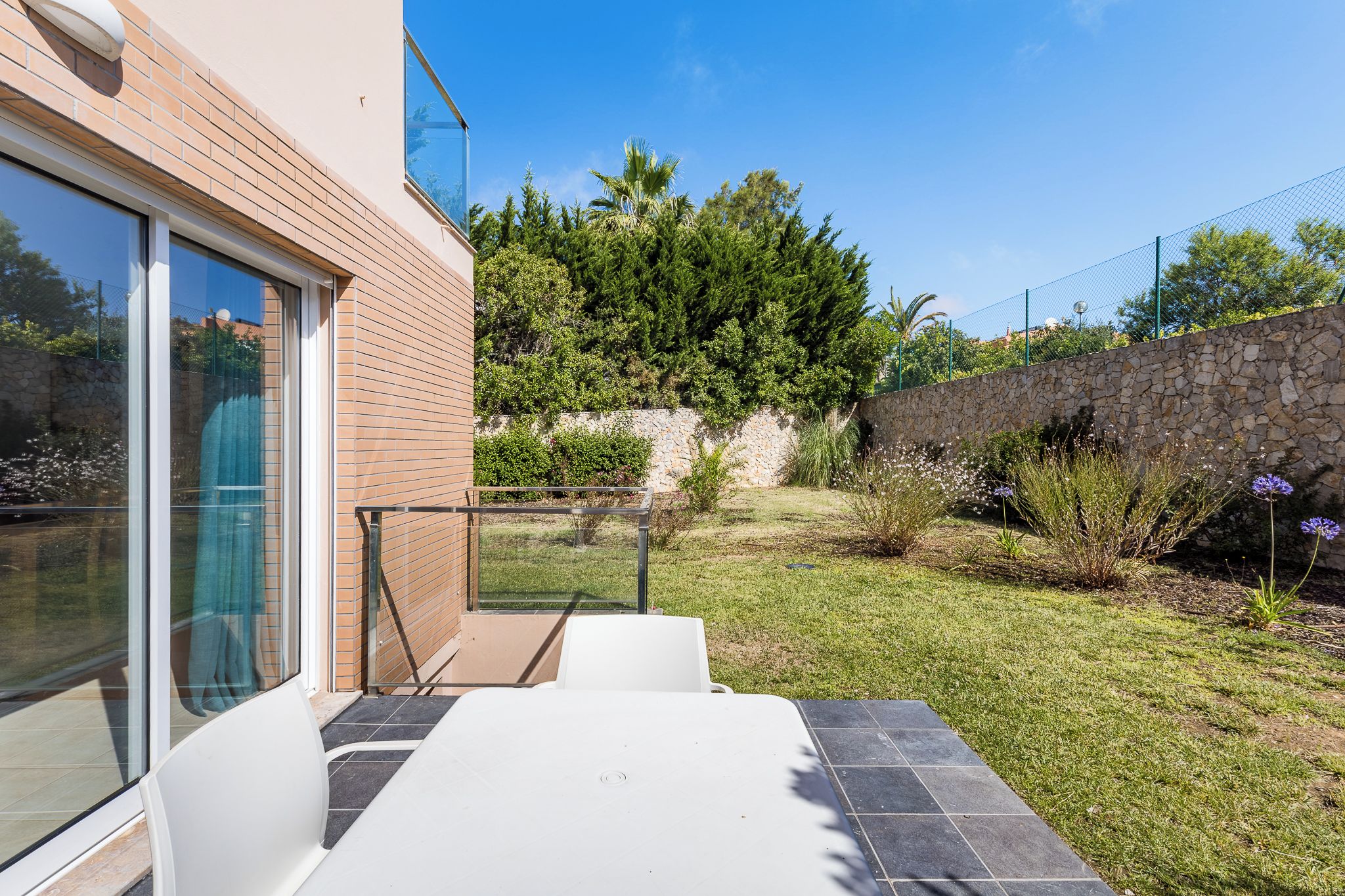 Eine Terrasse mit Tisch und Stühlen, eine Steinmauer und eine Rasenfläche mit Pflanzen und Blumen.