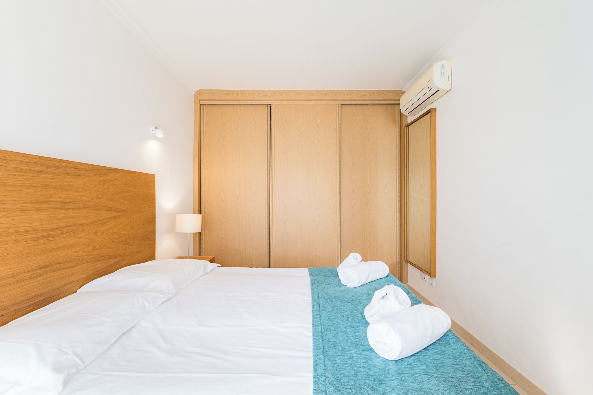 A hotel room with a wooden headboard, a bed with white linens, a teal throw blanket, and two rolled towels on the bed.