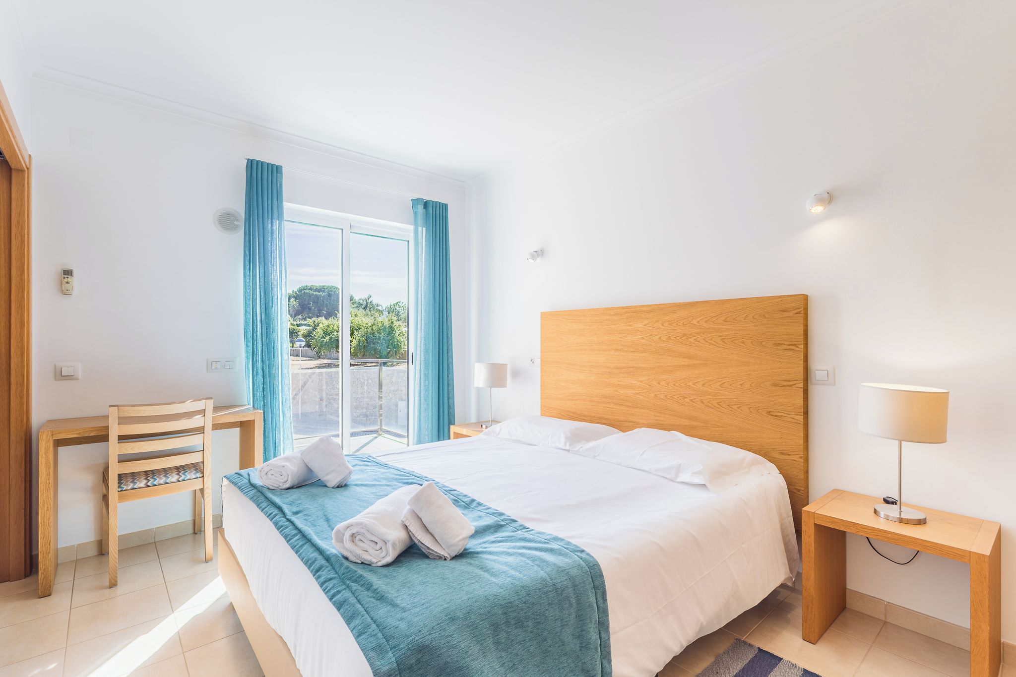 A hotel room with a bed, a desk, a chair, a nightstand, and a window with curtains.
